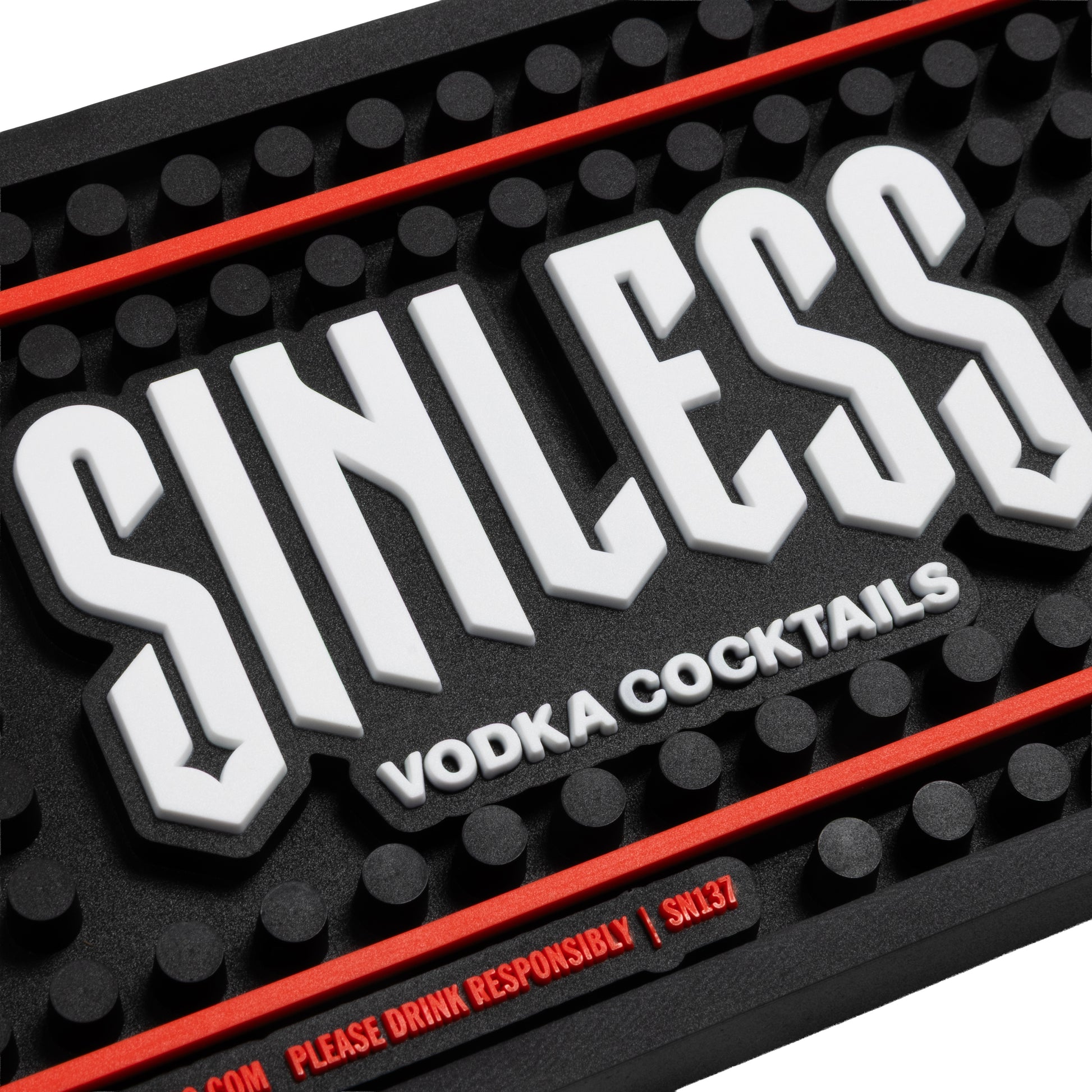 A close-up of a black textured bar mat with raised white 'SINLESS' lettering, smaller white 'VODKA COCKTAILS' text, and red accent lines and warning text.