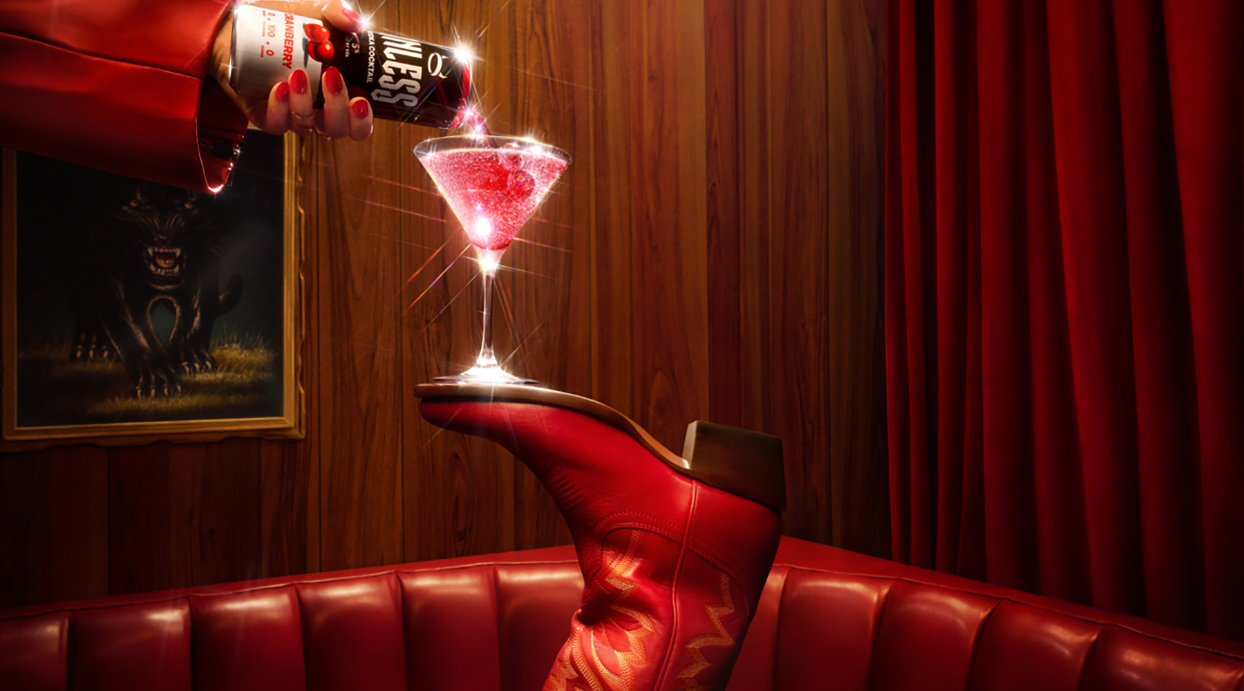 A hand in a red sleeve pours a Sinless Cranberry from a can into a martini glass balanced on a red leather boot, set in a dimly lit red leather booth with wood-paneled walls and red curtains.